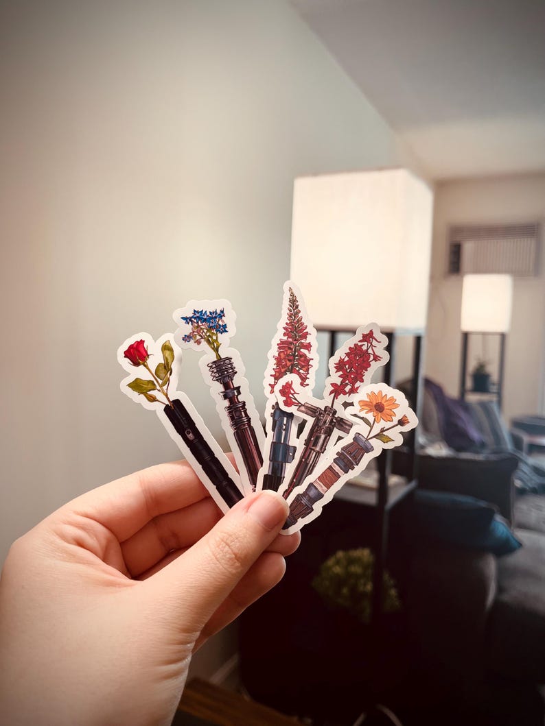 May include: Six different flower stickers with a lightsaber design. The flowers are a red rose, blue and white flowers, two red flowers, and a yellow sunflower.