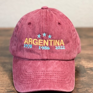 Puede incluir: Una gorra de béisbol de color granate con la palabra "ARGENTINA" bordada en amarillo con tres estrellas azules encima de la palabra. Los años "1978", "1986" y "2022" están bordados en azul debajo de la palabra.