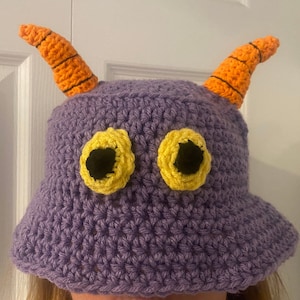Op de afbeelding: Een handgemaakte paarse gehaakte emmerhoed met oranje hoorns en gele ogen. De hoorns hebben zwarte strepen. De ogen zijn geel met zwarte centra. Deze grillige hoed is een leuk accessoire.
