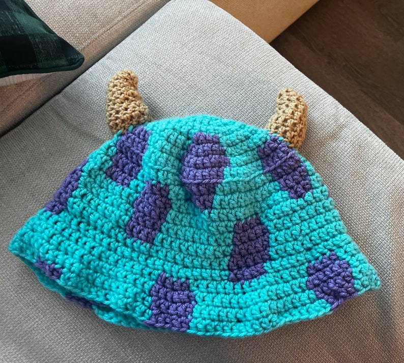 K&ouml;nnte beinhalten: Eine blaue und lila geh&auml;kelte M&uuml;tze mit zwei braunen H&ouml;rnern. Die M&uuml;tze ist in Form eines Bucket-Huts und hat ein strukturiertes Design.