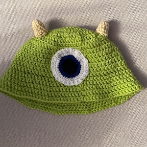 K&ouml;nnte beinhalten: Gr&uuml;ne H&auml;kelm&uuml;tze mit einem gro&szlig;en blauen Auge und wei&szlig;en und schwarzen Akzenten. Die M&uuml;tze hat zwei kleine beigefarbene H&ouml;rner oben.