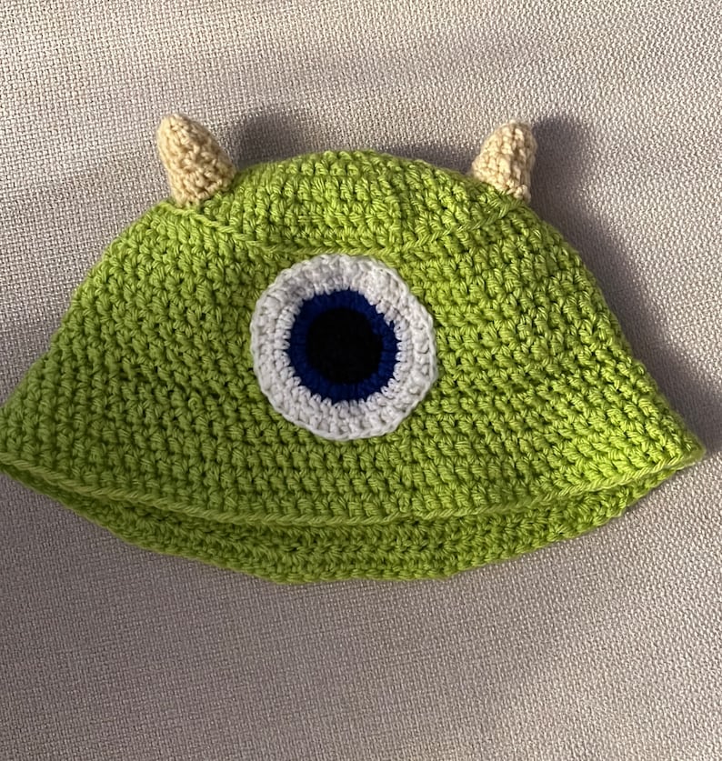 K&ouml;nnte beinhalten: Gr&uuml;ne H&auml;kelm&uuml;tze mit einem gro&szlig;en blauen Auge und wei&szlig;en und schwarzen Akzenten. Die M&uuml;tze hat zwei kleine beigefarbene H&ouml;rner oben.