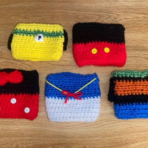 Op de afbeelding: Vijf gehaakte bekerwarmers in verschillende kleuren en ontwerpen. De bekerwarmers zijn gemaakt van verschillende kleuren garen en hebben verschillende ontwerpen, waaronder een gele en groene bekerwarmer met een letter "P", een rode en zwarte bekerwarmer met twee gele knopen, een blauwe en witte bekerwarmer met een rode strik, een zwarte en oranje bekerwarmer met groene strepen en een rode en zwarte bekerwarmer met een rode strik.