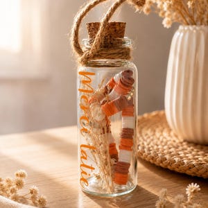 Puede incluir: Un tarro de cristal transparente con tapón de corcho y asa de cuerda, lleno de flores secas y cuentas decorativas. El tarro tiene la palabra "merci" en escritura naranja. Un jarrón blanco con flores secas está en el fondo.