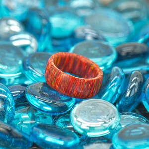 以下が含まれることがあります： 青いガラスの石のベッドの上に置かれた、自然な木目模様の赤いオレンジ色の木製のリングです。