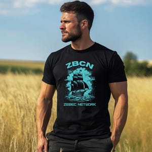 May include: Black t-shirt with a teal graphic of a ship at sea, the text "ZBCN" above the ship, and "ZEBEC NETWORK" below. The shirt is worn by a person standing in a field of tall grass under a blue sky.