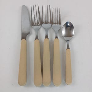 May include: A five-piece vintage flatware set. The set includes a knife, three forks, and a spoon. Each piece has a stainless steel head and a cream-colored handle.