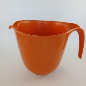 May include: An orange plastic mixing bowl with a handle. The bowl has a curved rim and a spout on one side. The handle is arched and attached to the side of the bowl. The bowl is empty and sits against a white background.