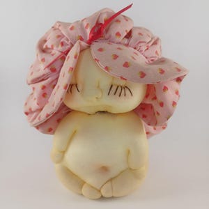 May include: A vintage doll with a light skin tone, wearing a pink bonnet adorned with small strawberry patterns. The doll's face features closed eyes and delicate painted details. The doll is in a seated position.