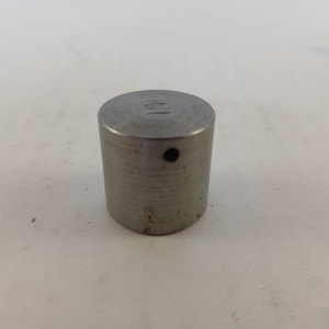 May include: A cylindrical metal object with a small hole on the side. The top surface has an engraved design. The object is made of a brushed metal material and has a slightly worn appearance.
