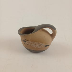 May include: A small, rounded ceramic pitcher with a tan body and dark gray accents on the rim, handle, and base. The exterior features a black and white painted feather design on the side, with a smooth, matte finish and a single curved handle.