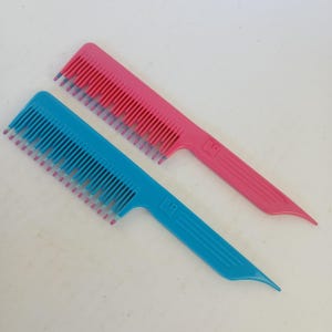 May include: Two plastic hair combs. One is bright pink, the other is turquoise. Both combs have a handle and fine teeth. The teeth of the combs are a lighter shade of pink and blue at the tips. The combs are on a white surface.