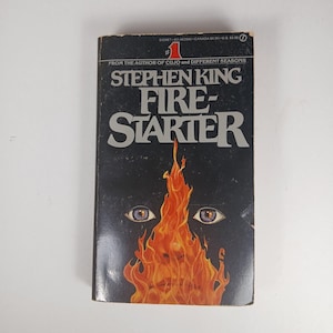 May include: A mass market paperback book titled Firestarter by Stephen King. The black cover features a large illustration of a face engulfed in bright orange and yellow flames, with the title in bold grey lettering and author name above.
