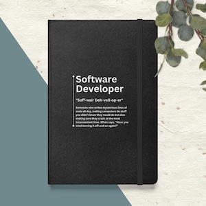 Puede incluir: Cuaderno negro con un título blanco que dice "Software Developer" y una definición del término. El cuaderno tiene una banda elástica negra.