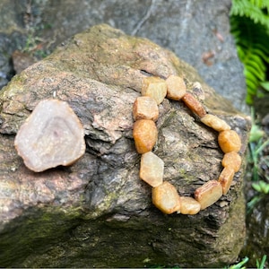Peut inclure: Un bracelet composé de perles de pierres précieuses brun clair et orange. Les perles sont facettées et ont une forme hexagonale. Le bracelet est posé sur un rocher avec une petite pierre précieuse ronde brun clair et blanche à côté.