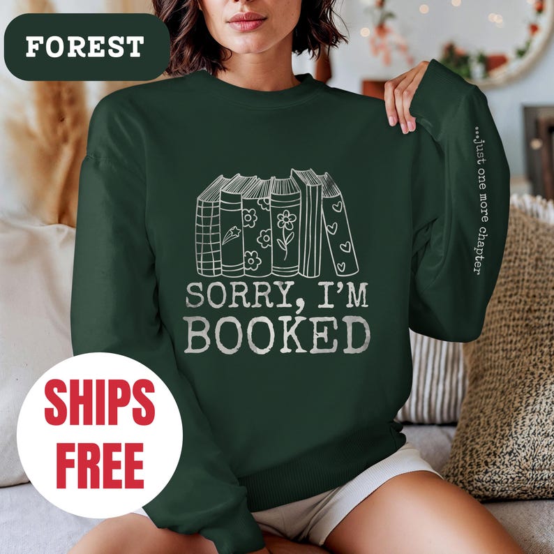 May include: A dark green sweatshirt with a white graphic of stacked books and the text "Sorry, I'm Booked." The text "...Just one more chapter" is printed on the sleeve.