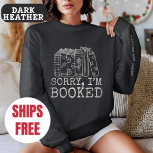 May include: Dark heather grey sweatshirt with a stack of books and the text "Sorry, I'm Booked" in white. The sleeve has the text "...just one more chapter" in white.