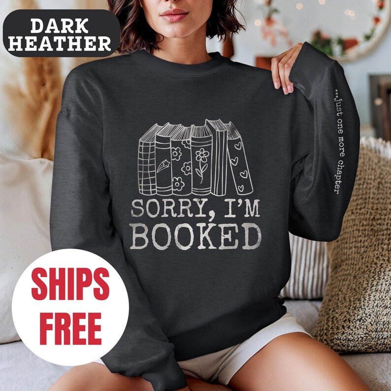 May include: Dark heather grey sweatshirt with a stack of books and the text "Sorry, I'm Booked" in white. The sleeve has the text "...just one more chapter" in white.