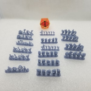May include: A set of 12 miniature grey plastic figures, arranged in rows on a white surface. A red and gold dice is in the center of the image.