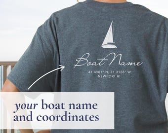 May include: Dark gray t-shirt with a white sailboat graphic and the words "Boat Name" in script. Below the text are coordinates and the city "Newport RI". The shirt also has the text "your boat name and coordinates".