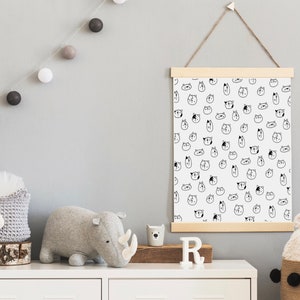 May include: A white canvas print with a black and white pattern of animal faces. The print is hanging on a wall with a wooden frame and a string. There are gray and white cotton ball lights hanging on the wall. A gray stuffed rhinoceros and a gray stuffed animal with a white face are on a white dresser with a wooden top. There is a white letter "R" on the dresser.