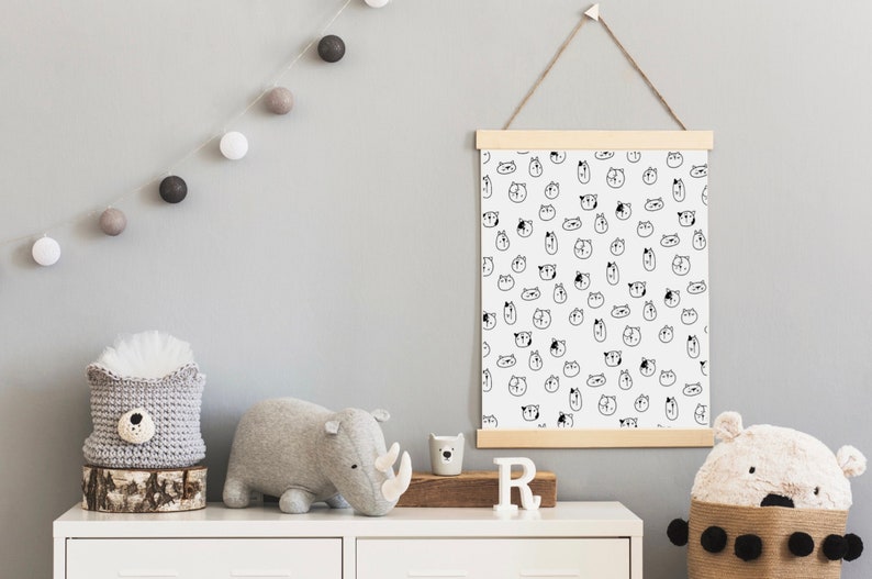 May include: A white canvas print with a black and white pattern of animal faces. The print is hanging on a wall with a wooden frame and a string. There are gray and white cotton ball lights hanging on the wall. A gray stuffed rhinoceros and a gray stuffed animal with a white face are on a white dresser with a wooden top. There is a white letter "R" on the dresser.