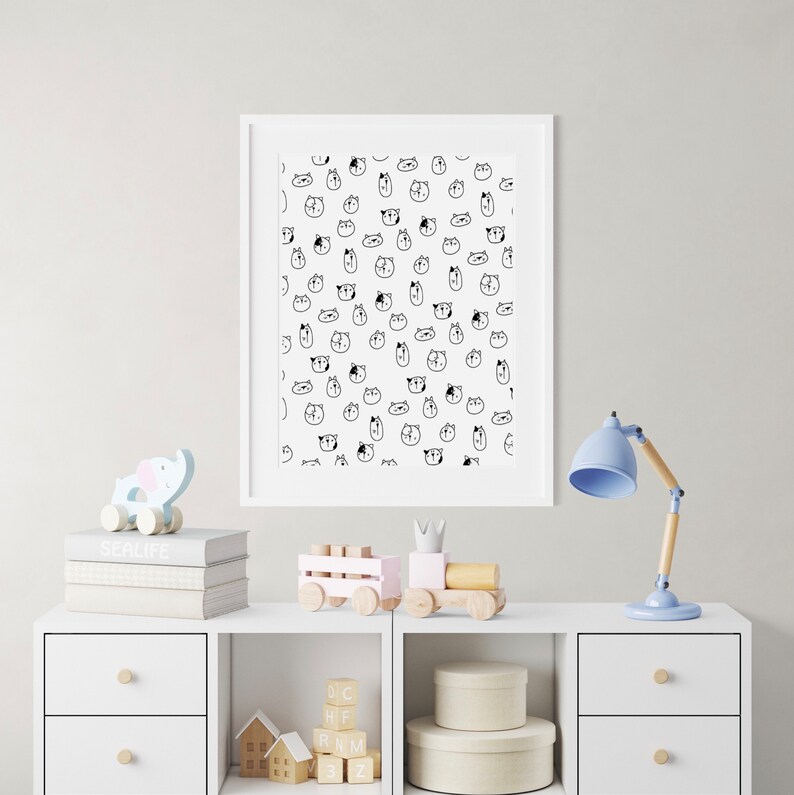 May include: A white framed print with a black and white pattern of cartoon animal faces. The print is hanging on a white wall above a white dresser with a wooden toy train, a wooden elephant toy, and a blue lamp.