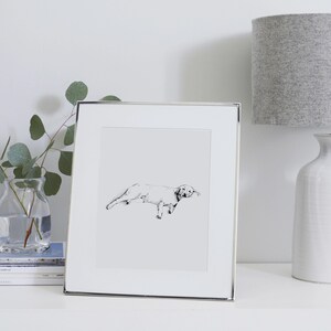 K&ouml;nnte beinhalten: Gerahmte Schwarz-Wei&szlig;-Skizze eines Hundes. Das Kunstwerk befindet sich in einem silbernen Rahmen und wird auf einer wei&szlig;en Oberfl&auml;che pr&auml;sentiert. Eine graue Lampe und eine Vase mit Gr&uuml;n sind ebenfalls zu sehen.