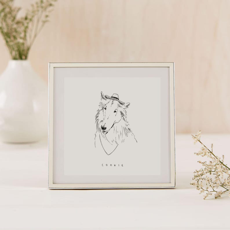 K&ouml;nnte beinhalten: Ein gerahmter Kunstdruck mit einer Strichzeichnung eines Hundes mit Hut und Schal. Der Name des Hundes, "COOKIE", steht unter dem Bild. Der Rahmen ist silberfarben, der Hintergrund ist neutral.