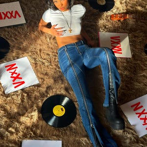 Pu&ograve; includere: Una donna indossa un top bianco corto e jeans blu con cerniere sui lati. &Egrave; sdraiata su un tappeto di pelo marrone con un giradischi e una chitarra sullo sfondo. Ci sono diversi pezzi di carta quadrati con la parola "VXXN" stampata in rosso.