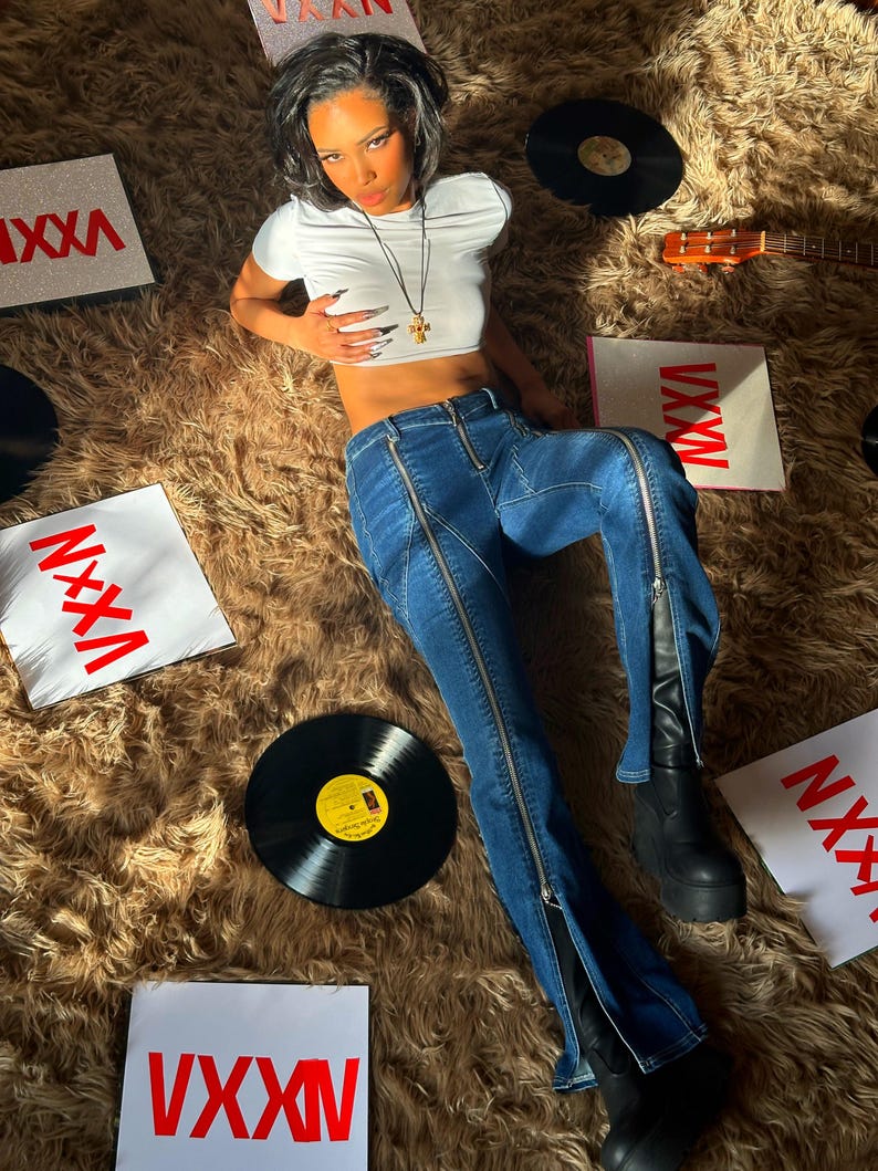 Pu&ograve; includere: Una donna indossa un top bianco corto e jeans blu con cerniere sui lati. &Egrave; sdraiata su un tappeto di pelo marrone con un giradischi e una chitarra sullo sfondo. Ci sono diversi pezzi di carta quadrati con la parola "VXXN" stampata in rosso.