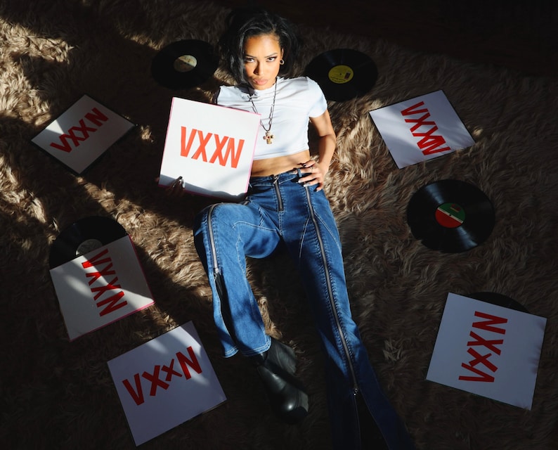 Pu&ograve; includere: Una donna con un top bianco corto e jeans blu posa su un tappeto peloso con diversi dischi in vinile e quadrati bianchi con la parola "VXXN" stampata in rosso.