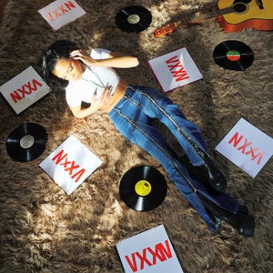 Pu&ograve; includere: Una donna con una camicia bianca e jeans blu con cerniere sui lati &egrave; sdraiata su un tappeto di pelo marrone. Indossa delle cuffie e ascolta musica. Ci sono diversi dischi in vinile e quadrati bianchi con il testo "VXXN" sparsi intorno a lei. Una chitarra &egrave; sdraiata sul tappeto.