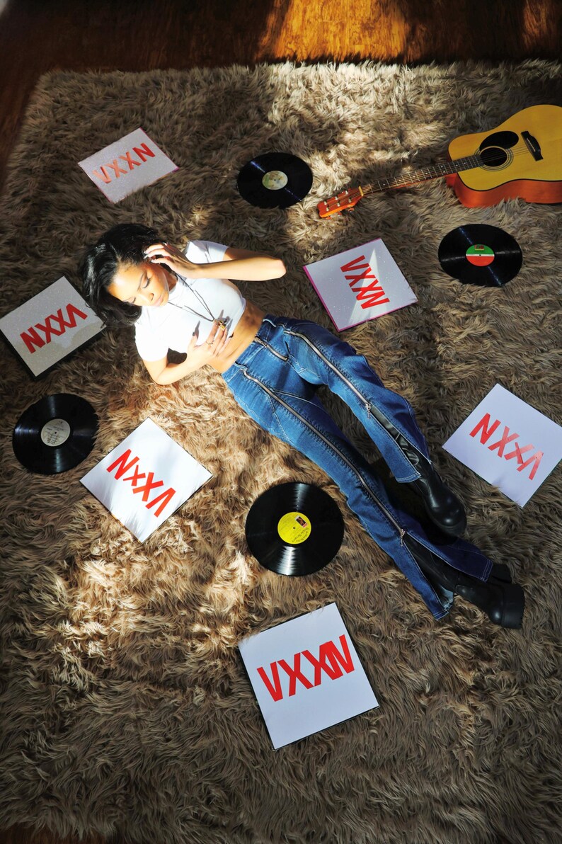 Pu&ograve; includere: Una donna con una camicia bianca e jeans blu con cerniere sui lati &egrave; sdraiata su un tappeto di pelo marrone. Indossa delle cuffie e ascolta musica. Ci sono diversi dischi in vinile e quadrati bianchi con il testo "VXXN" sparsi intorno a lei. Una chitarra &egrave; sdraiata sul tappeto.