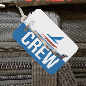 May include: A black luggage bag with a red "CREW" strap and a white luggage tag with a blue background and the text "CREW" and the Piedmont Airlines logo.