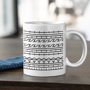 May include: White ceramic mug with a black geometric pattern. The design features horizontal lines, triangles, and other shapes. The mug has a curved handle and is sitting on a wooden surface.