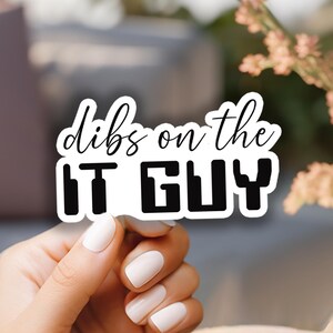 May include: A white sticker with black text that reads "dibs on the IT GUY". The sticker is held by a hand with white nail polish. The background is blurred, with a vase of pink flowers.
