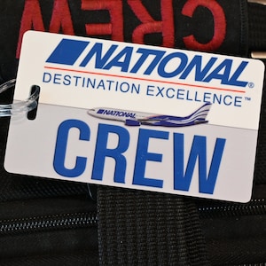 May include: A white and blue luggage tag with the text "NATIONAL DESTINATION EXCELLENCE" and "CREW". The tag features a small airplane graphic and is attached to a black bag.