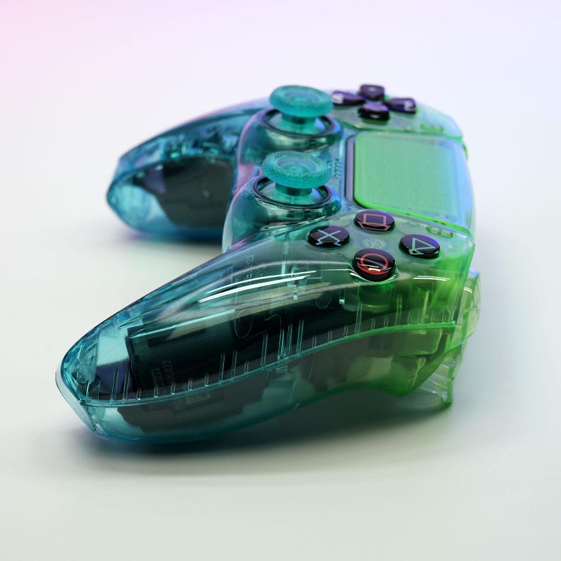 Peut inclure: Une manette de jeu vid&eacute;o transparente, de couleur vert d'eau et verte, avec des boutons et des joysticks noirs. La manette a un design clair et transparent, mettant en valeur les composants internes. Les boutons incluent des fl&egrave;ches directionnelles, un carr&eacute;, un X, un cercle et un triangle.