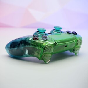 Peut inclure: Une manette de jeu vid&eacute;o verte et bleue transparente. La manette a une coque en plastique transparent, avec des accents verts et bleus sur les boutons et les joysticks. Les boutons sont noirs et rouges. La manette est con&ccedil;ue pour le jeu.