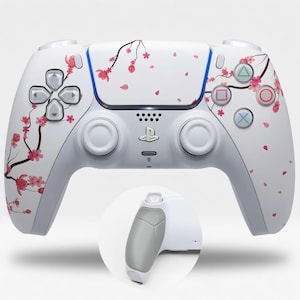 Puede incluir: Un mando de juego inalámbrico blanco con un patrón de ramas de cerezo en flor de color rosa en la carcasa frontal. El mando incluye dos joysticks, una cruceta, cuatro botones de acción y un logotipo de PlayStation central. Un recuadro circular muestra la empuñadura gris texturizada.