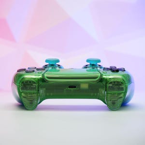 Peut inclure: Une manette de jeu verte transparente avec des joysticks bleus et des boutons noirs. La manette a un design &eacute;l&eacute;gant et moderne avec une coque transparente, permettant de voir les composants internes. L'arri&egrave;re-plan est un d&eacute;grad&eacute; de rose et de violet.