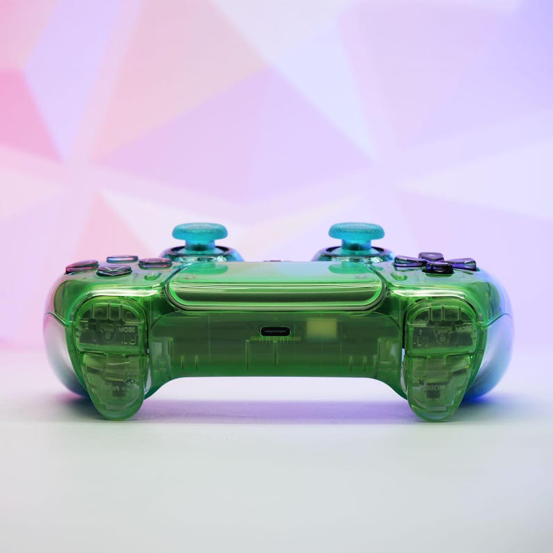 Peut inclure: Une manette de jeu verte transparente avec des joysticks bleus et des boutons noirs. La manette a un design &eacute;l&eacute;gant et moderne avec une coque transparente, permettant de voir les composants internes. L'arri&egrave;re-plan est un d&eacute;grad&eacute; de rose et de violet.