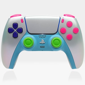 Puede incluir: Un mando personalizado de PlayStation 5 con centro plateado, detalles en azul y botones rosas y verdes. El mando presenta una cruceta con botones morados y el logotipo de PlayStation.