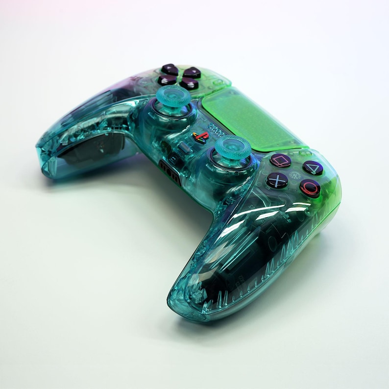 Peut inclure: Une manette de jeu vid&eacute;o transparente, de couleur vert d'eau et verte, avec des boutons noirs et un pav&eacute; tactile vert. La manette a un design clair, laissant voir les composants internes. Le logo PlayStation est visible.