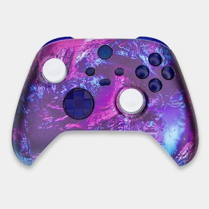 Puede incluir: Un mando de videojuegos inalámbrico con un vibrante diseño veteado en morado, azul y rosa. El mando presenta palancas blancas, una cruceta azul y varios botones negros. El borde superior es de color azul liso.