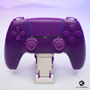 May include: A purple PlayStation 5 DualSense controller with a white stand. The controller has a purple faceplate and purple buttons.