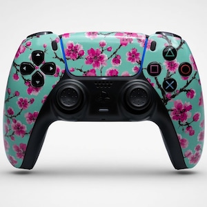 Puede incluir: Un mando de videojuegos inalámbrico con una base de color verde azulado claro y un patrón repetitivo de flores de cerezo rosas y ramas de color marrón oscuro. El mando incluye joysticks negros, una cruceta negra y botones de acción negros.