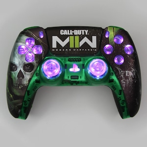 May include: A green and black PlayStation 5 controller with a skull design and the words "Call of Duty: Modern Warfare II" on the front. The controller has purple lights on the buttons and triggers.