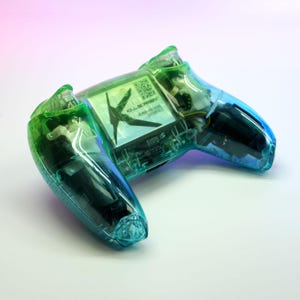 Peut inclure: Une manette de jeu vid&eacute;o transparente, de couleur vert d'eau et verte, avec des composants internes visibles. La manette pr&eacute;sente un d&eacute;grad&eacute; de couleurs et le texte "KILLSCREEN" et "HUMAN-MACHINE INTERFACE".